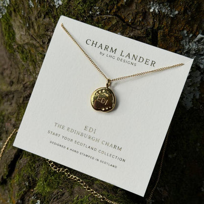 A gold-toned charm necklace displayed on a white branded card, placed against a mossy tree trunk. The card reads 'CHARM LANDER by LHG DESIGNS' and 'EDI THE EDINBURGH CHARM'.