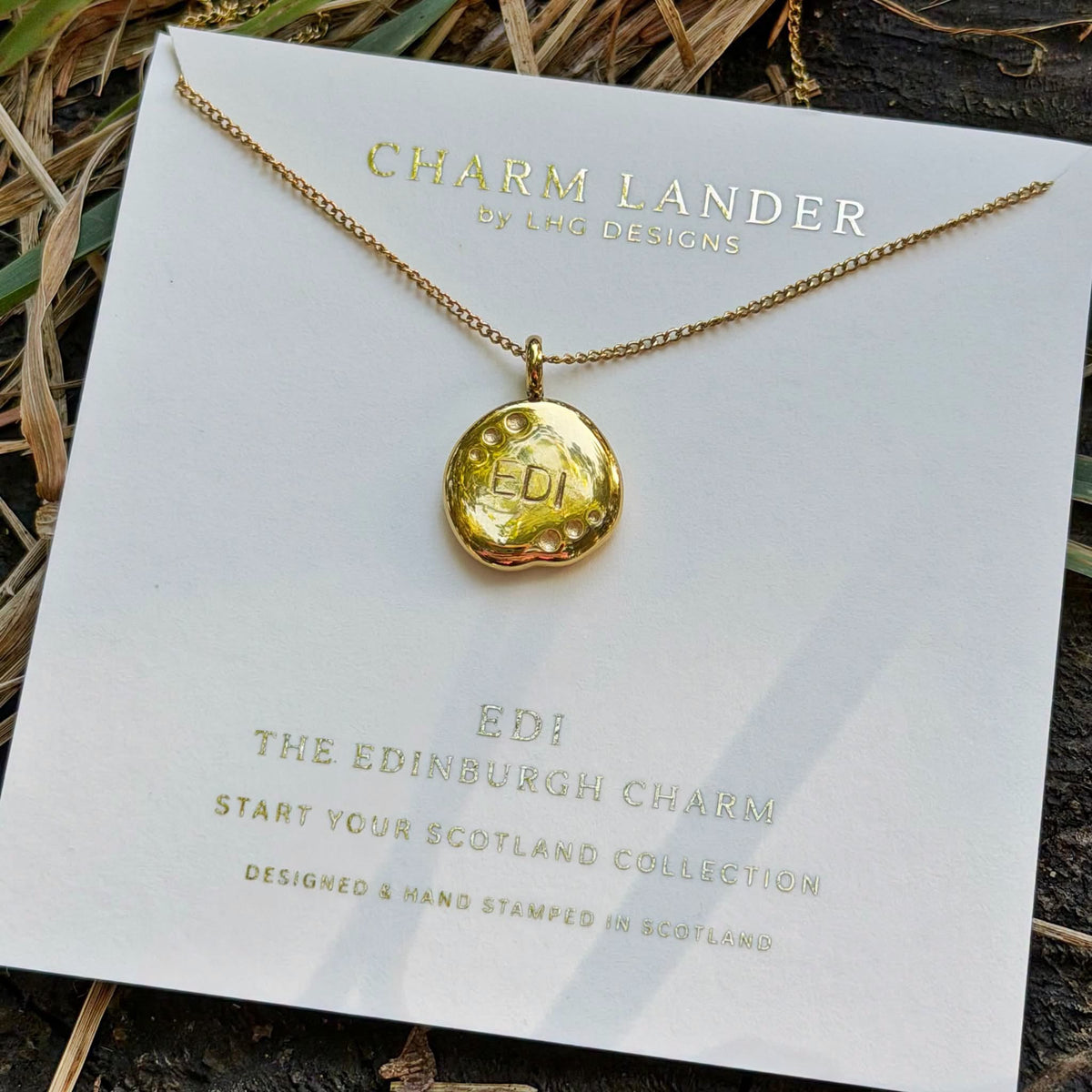 A gold necklace with a round stamped charm reading 'EDI' is displayed on a white branded card that says 'Charm Lander by LHG Designs.' The card is photographed outdoors on a dark textured surface with dried grass and greenery around it.