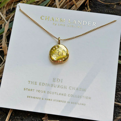A gold necklace with a round stamped charm reading 'EDI' is displayed on a white branded card that says 'Charm Lander by LHG Designs.' The card is photographed outdoors on a dark textured surface with dried grass and greenery around it.